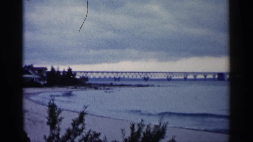 1960 Michigan 8mm frame with blue-tinted water, gentle surf, a distant bridge, left-side trees, and visible film dust.