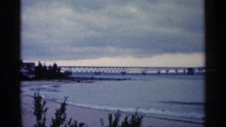 1960 Michigan beach view with foreground shrubs, pale sand, gentle waves, and a long bridge receding across the water under clouds.