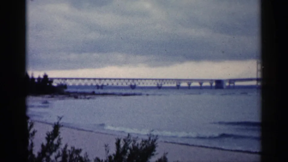1960 Michigan home-movie frame with a long truss and suspension bridge viewed across calm water from the shore.