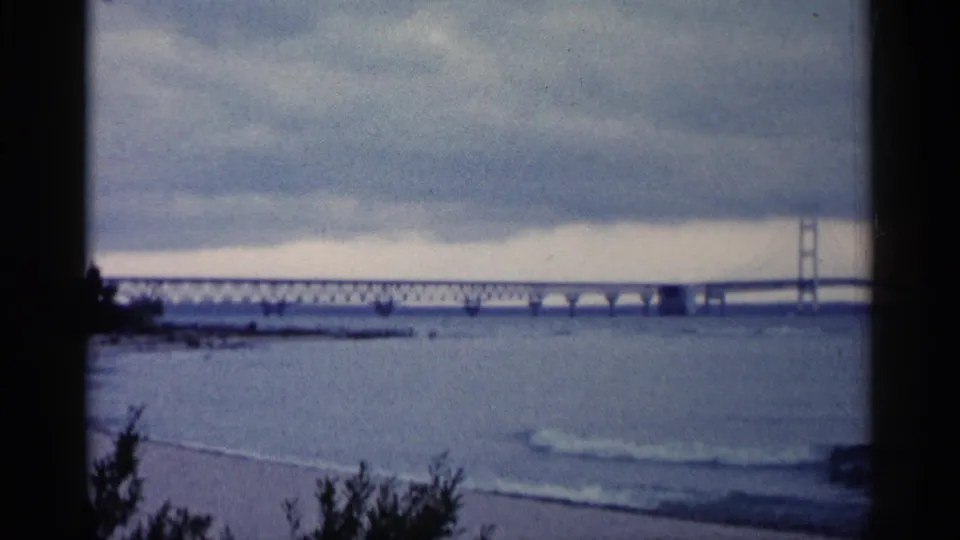 1960 Michigan 8mm frame showing the bridge tower and truss line across water, with beach edge and dark foliage below.