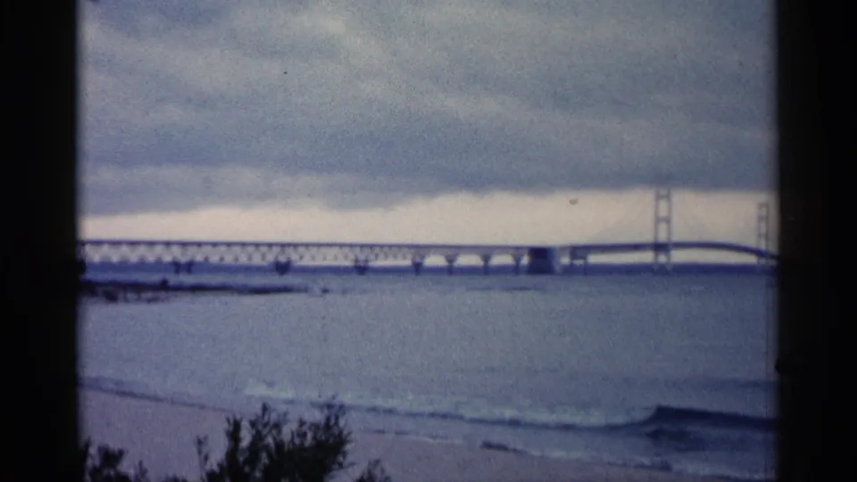 1960 Michigan home-movie frame of a long bridge crossing open water, with small waves and dark plants along the lower edge.