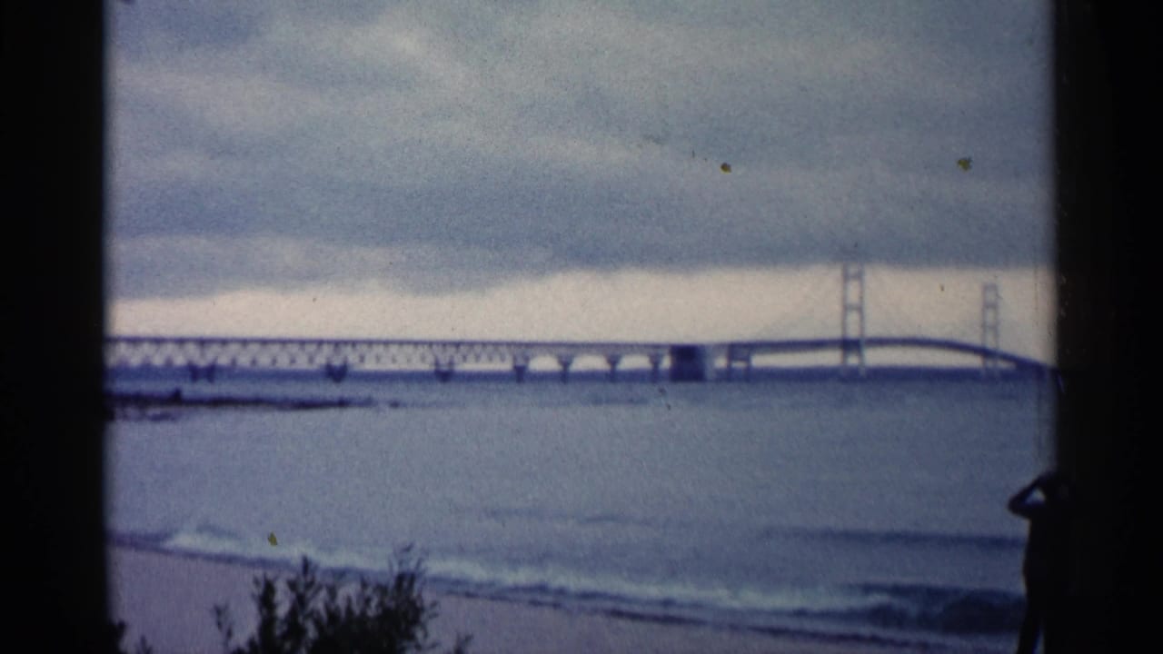 1960 Michigan 8mm frame showing a distant bridge across open water, small waves, beach plants, and a silhouetted man at the shoreline.