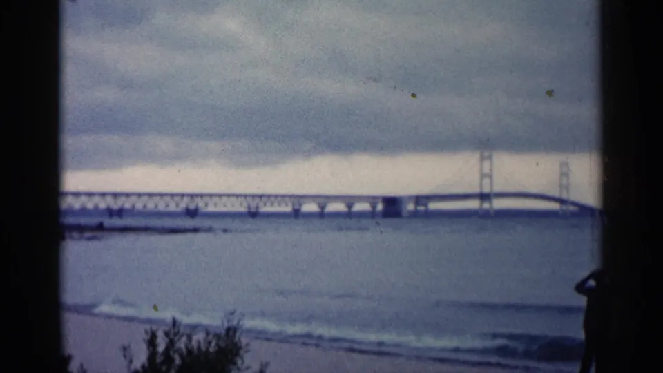 1960 Michigan 8mm frame showing a distant bridge across open water, small waves, beach plants, and a silhouetted man at the shoreline.