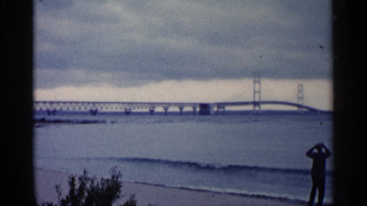 1960 Michigan shoreline view with a silhouetted man using binoculars toward a large bridge across a wide body of water.