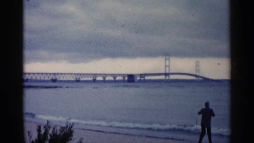 A 1960 Michigan home-movie frame shows a bridge beyond the water and a silhouetted beachgoer standing near gentle surf.