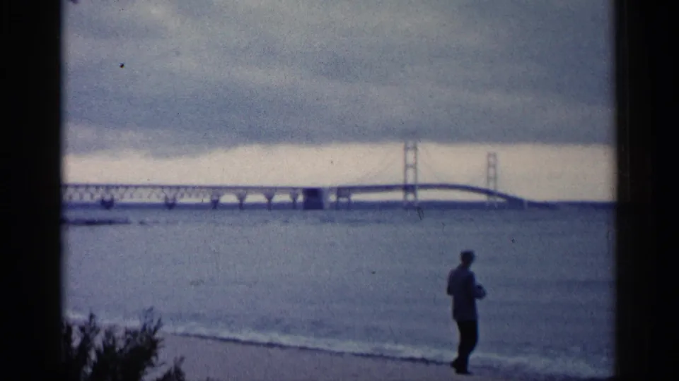 Final 1960 Michigan shoreline frame with a silhouetted man near the water and a large bridge visible through soft 8mm focus.