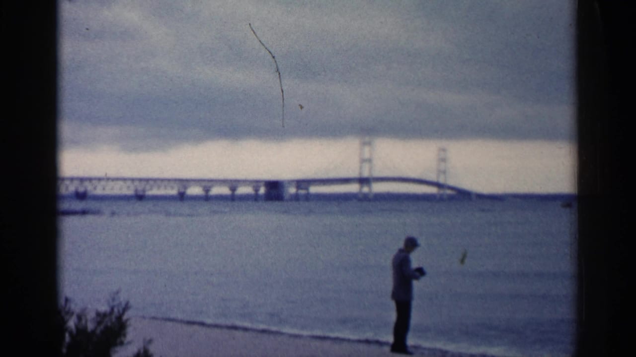 Suspension bridge over water with a lone shoreline figure under an overcast sky.