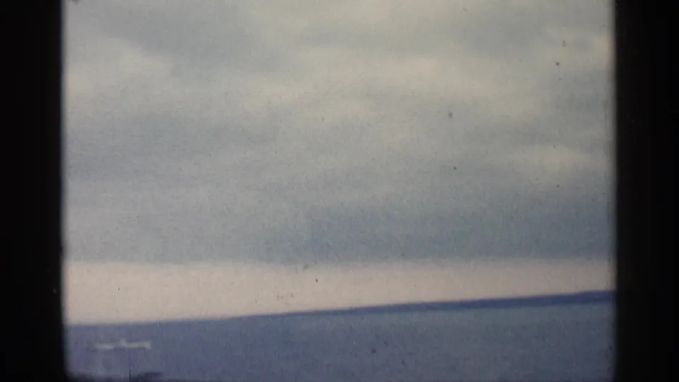 1960 Michigan waterfront view from 8mm footage, with distant shore, overcast sky, and a small pale shape on the water.