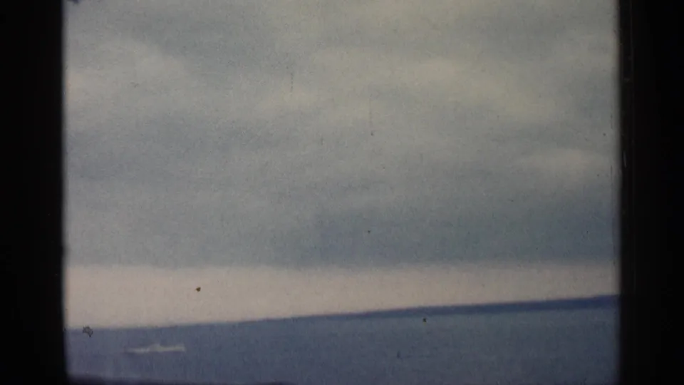 1960 Michigan 8mm waterfront frame with a low horizon, heavy clouds, and a small pale shape crossing the water.