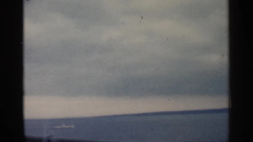 1960 Michigan home-movie frame showing a cloudy waterfront through dark window or railing divisions, with water and a distant shore.