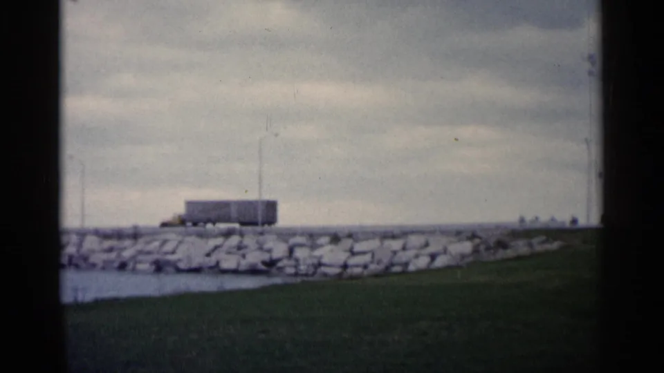 1960 Michigan shoreline scene with stone embankment, grassy foreground, small utility-like structure, poles, water, and cloudy sky.