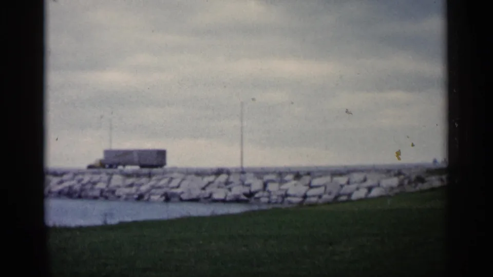 1960 Michigan home-movie frame showing a stone-lined waterfront, grass, small structure, pole, water edge, and overcast sky.