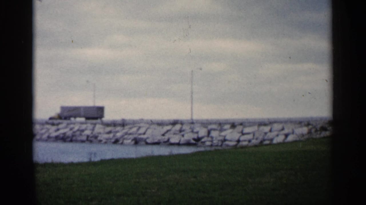 Distant semi-truck moving along a stone shoreline with water and grass in front.