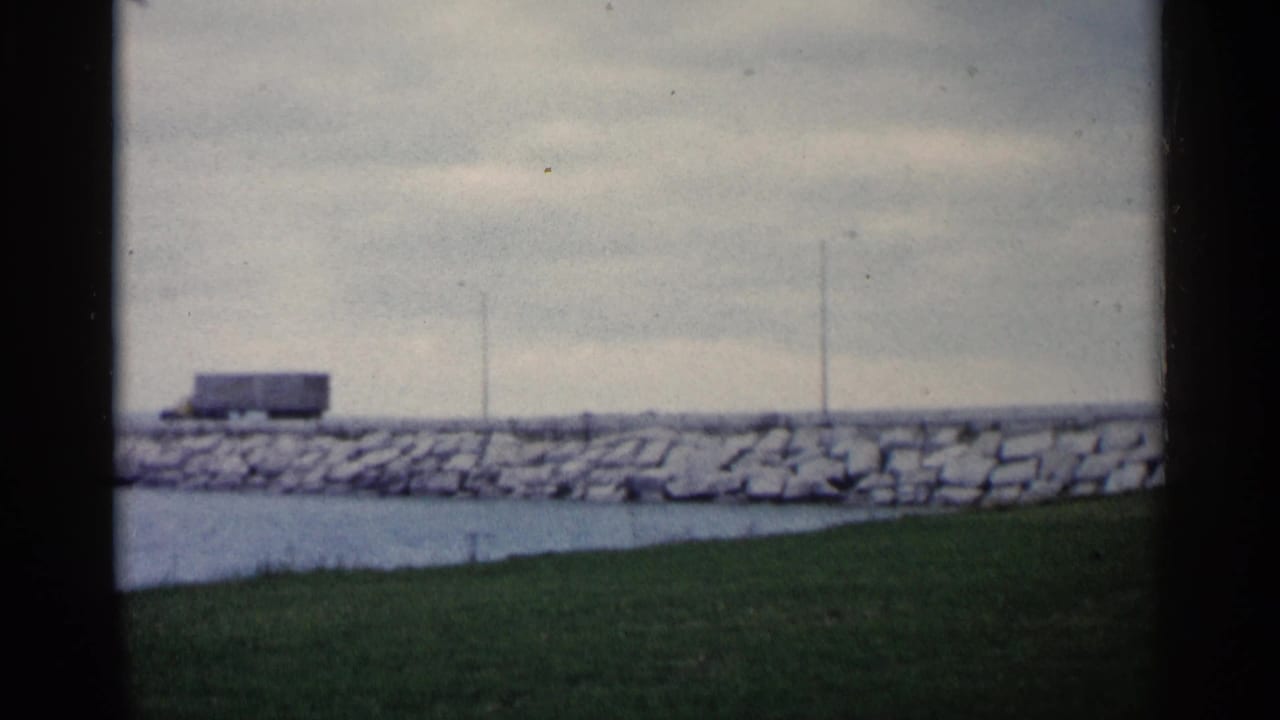 The truck appears farther along the Michigan shoreline road, framed by rocks, water, grass, and gray sky.