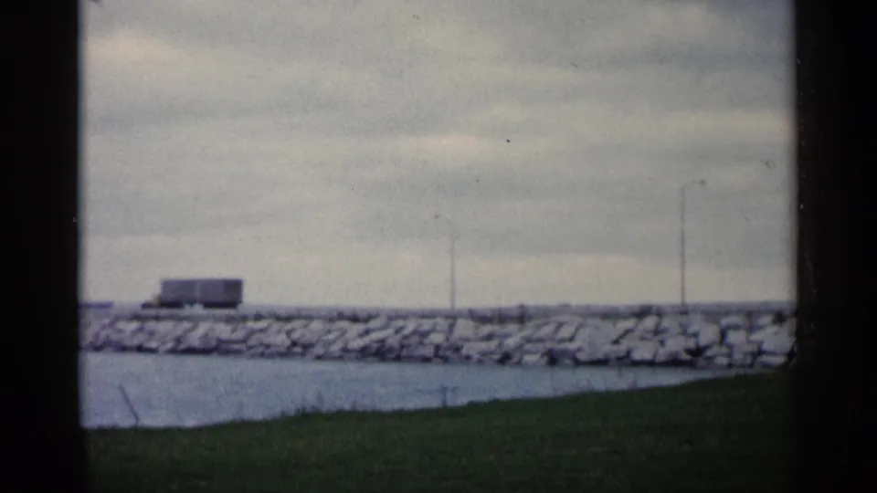 A soft 8mm view of the semi-truck progressing along the stone shoreline in Michigan, with water and grass below.