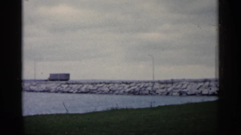 The semi-truck nears the right edge of the 1960 Michigan shoreline view, passing above calm water and grass.