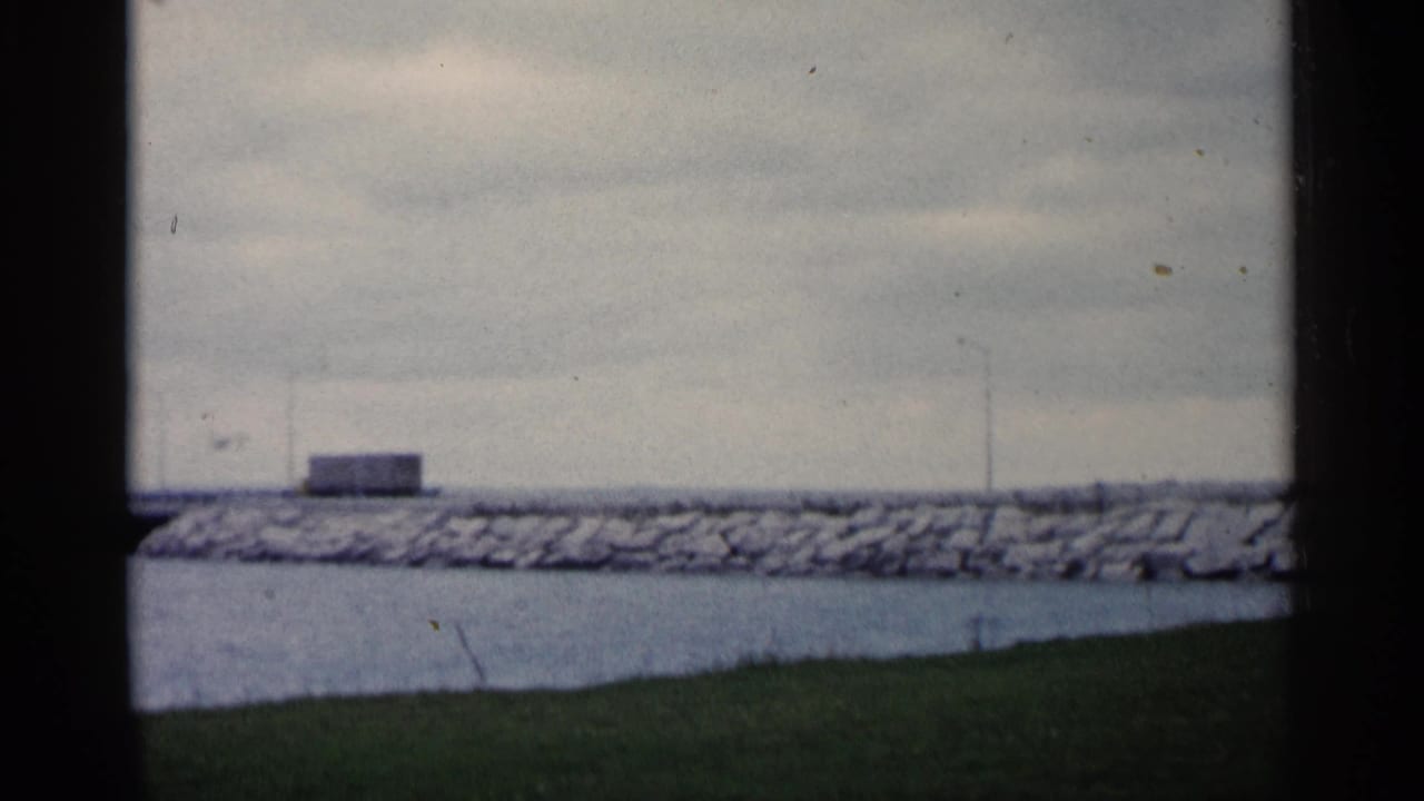1960 Michigan 8mm frame showing a distant bridge or causeway over water, with a rocky breakwater, grass, and an overcast sky.