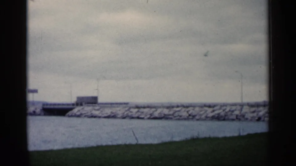1960 Michigan home-movie frame with a bridge or causeway crossing water beside a rocky breakwater and low shoreline structures.