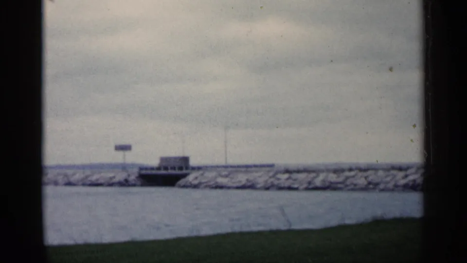 1960 Michigan 8mm frame showing a distant truck-like vehicle on a bridge, with water, breakwater rocks, grass, and a small sign visible.