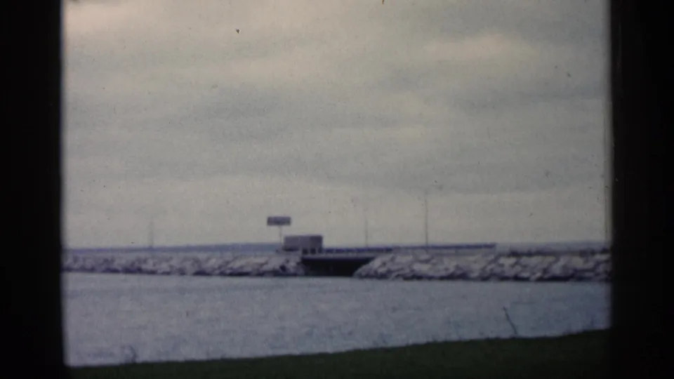 1960 Michigan home-movie frame of a distant vehicle crossing a bridge over water, framed by grass, breakwater rocks, and a cloudy sky.