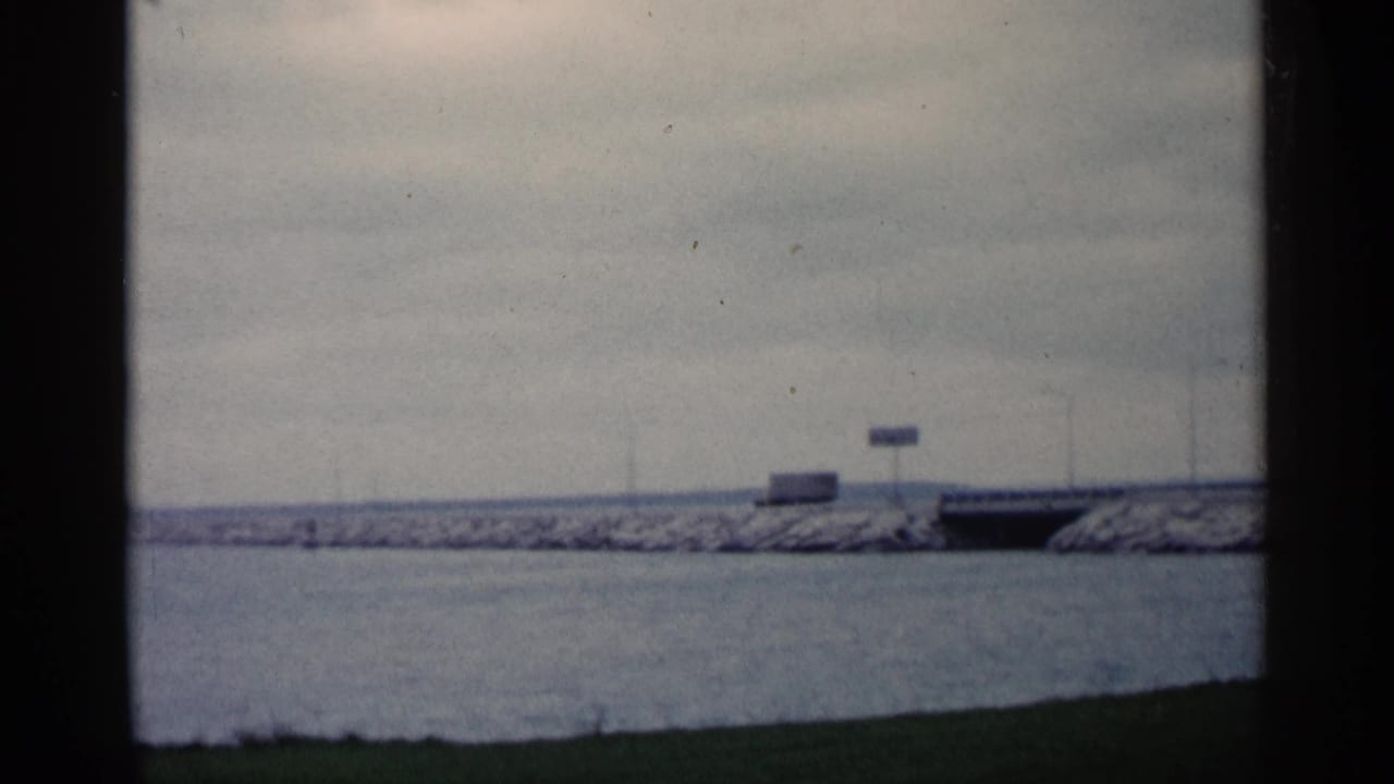 Long breakwater across calm water with a dark opening and signs near the far shore.