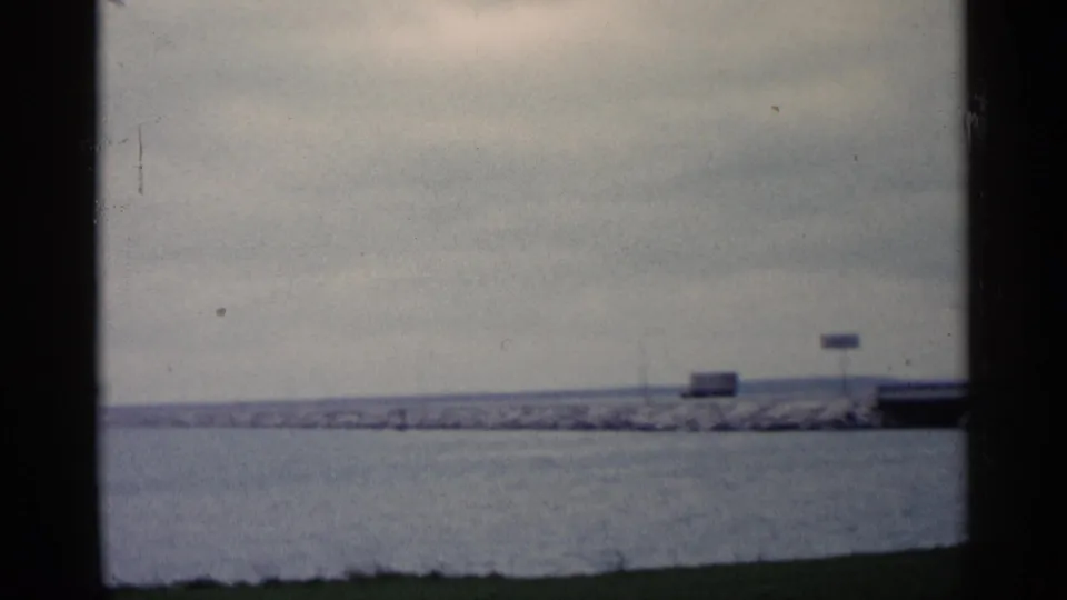 Grainy 8mm view of a Michigan breakwater, open water, small signs, and cloudy sky.