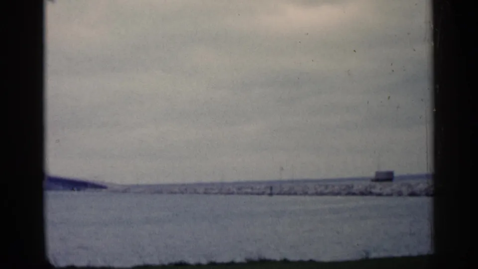 Wide 1960 Michigan shoreline frame with muted water, low breakwater, cloudy sky, and film borders.