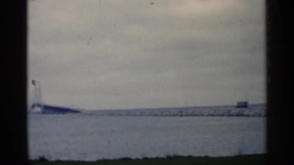 1960 Michigan home-movie frame showing a long bridge over a wide body of water beneath a gray sky.
