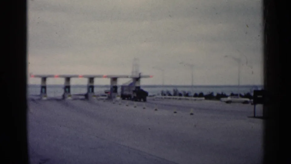 1960 Michigan highway approach toward a toll plaza, with vehicles, lane markers, and red signal lights visible.