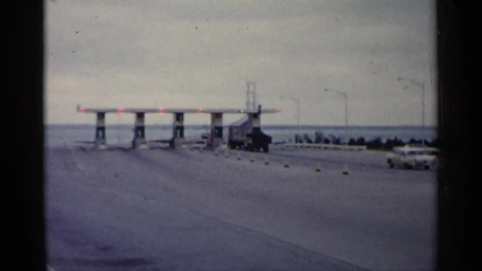 Home-movie view of a Michigan toll plaza near a bridge approach, with a truck, car, lane markers, and overcast sky.