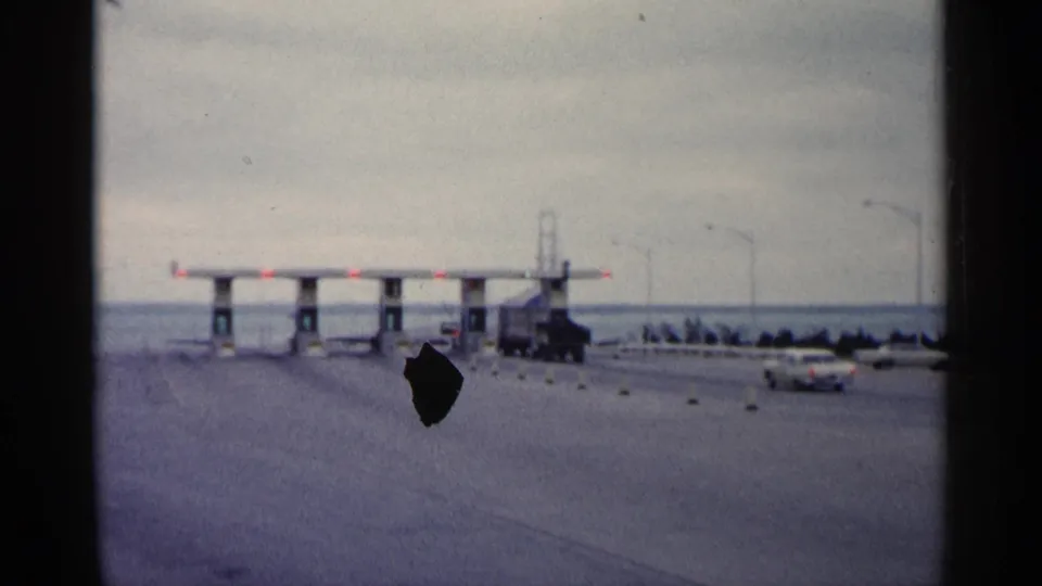 1960 Michigan 8mm frame showing a car driving toward ferry entrance gates, with a truck near the booths and water beyond.
