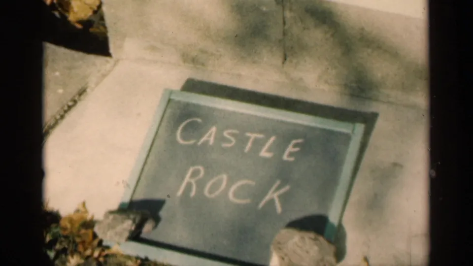 Handheld 8mm view of a chalkboard sign reading Castle Rock on pavement, with sidewalk and leaves around it.