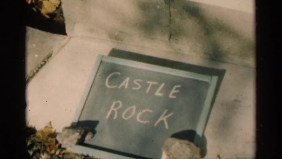 1960 Michigan home-movie frame with a tilted Castle Rock chalkboard sign on pavement near leaves and a concrete sidewalk.