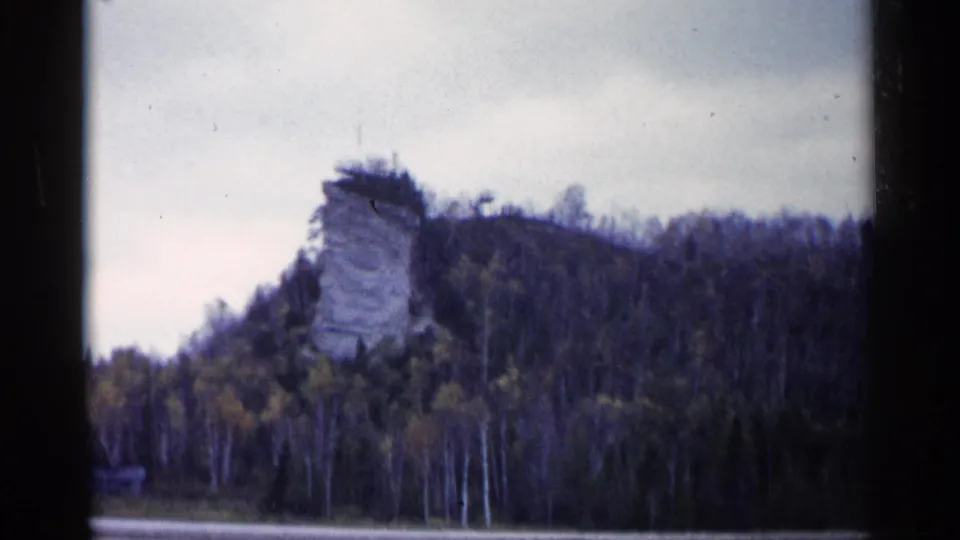 Soft 8mm frame of Castle Rock above a wooded slope, with autumn trees and water visible below.