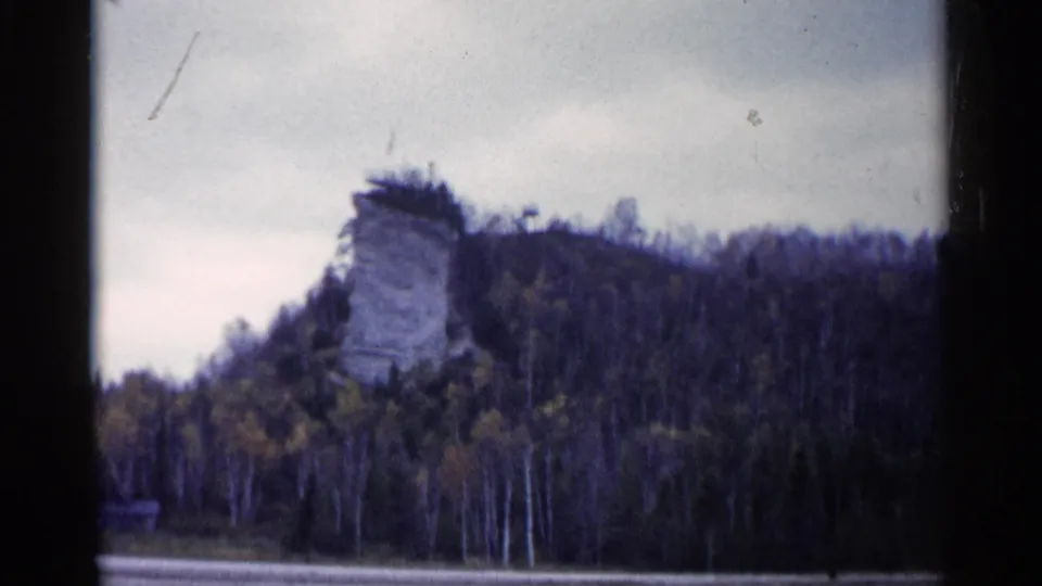 1960 Michigan 8mm view of Castle Rock on a wooded hillside, framed by sky, water, and dark film edges.