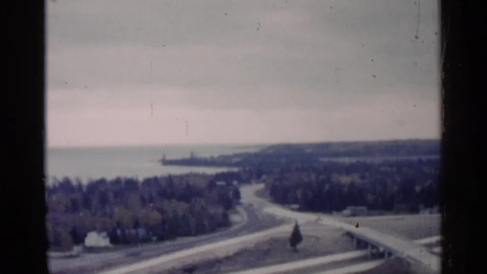 1960 Michigan 8mm frame with a broad overlook across roadways, forest, open land, and distant lake shoreline.
