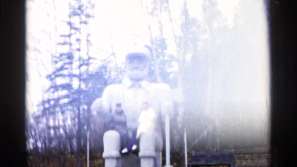 Overexposed 1960 Michigan frame of two adults seated on a large bearded statue in a wooded setting.