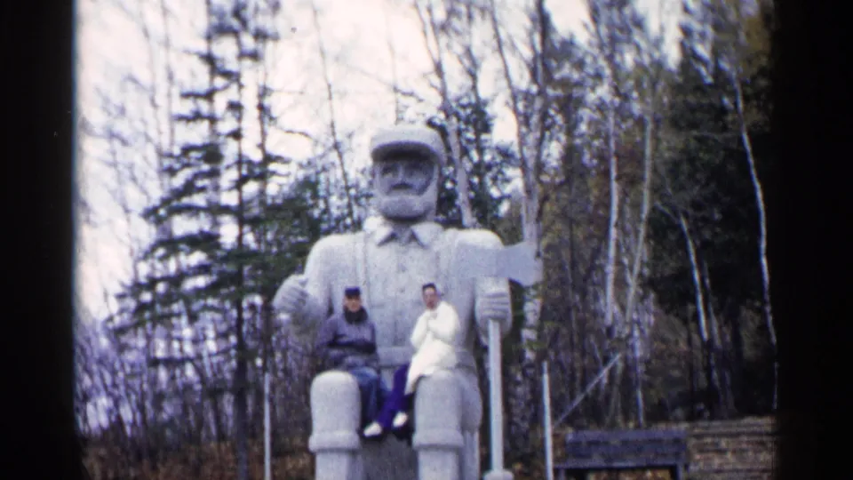 1960 Michigan home-movie frame of a couple posing on the footrest of a large bearded statue among autumn trees.