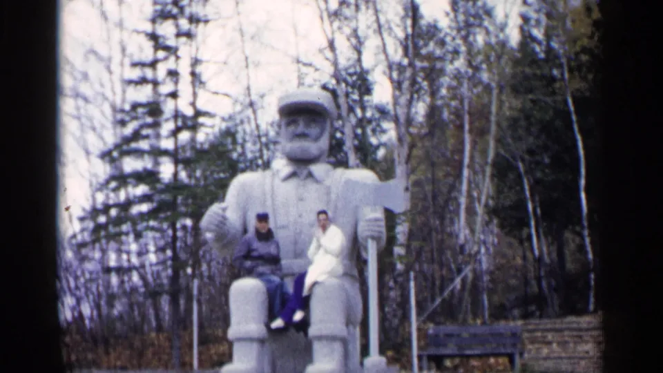 1960 Michigan 8mm frame showing two adults seated on a large bearded statue, with railings, bench, and woods behind.