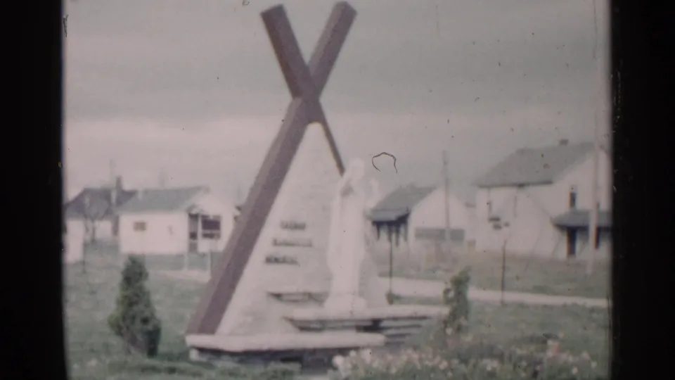 1960 Michigan outdoor monument with a large cross-like form, a robed statue, lawn, flowers, and small houses in the background.