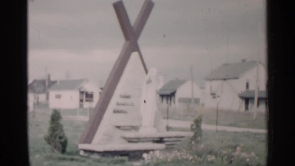 Another 1960 Michigan frame of the cross-shaped monument and statue, with modest houses and lawn visible behind it.