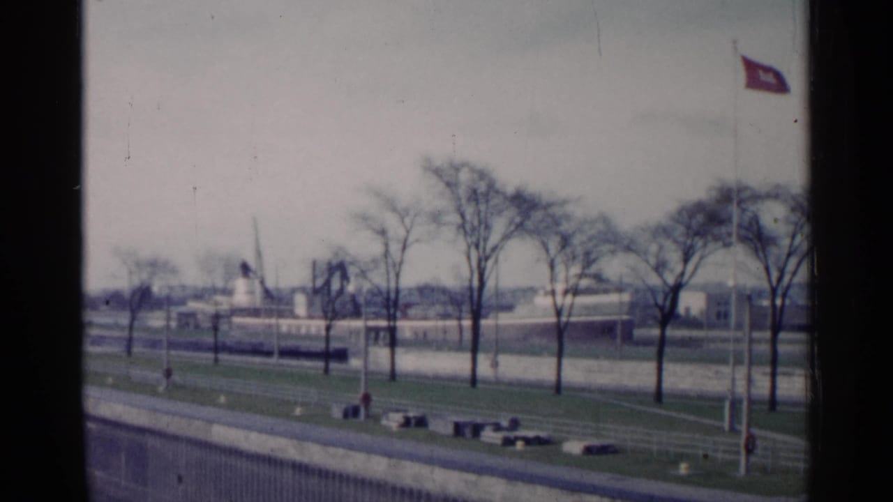 1960 Michigan 8mm frame showing bare trees, paved paths, a grassy embankment, distant waterfront buildings, and a red flag.