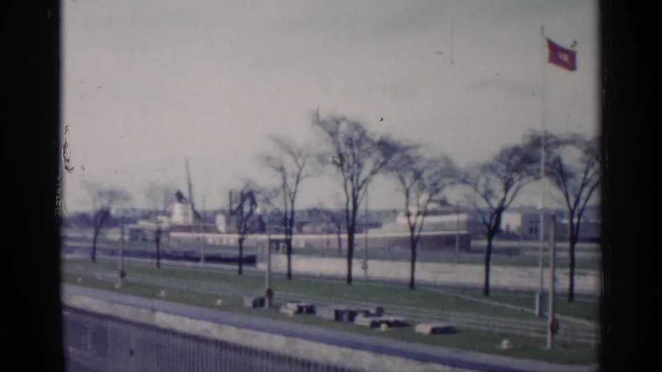 A soft 8mm view of a Michigan waterfront in 1960, with leafless trees, walkways, distant structures, and a red flag flying at right.