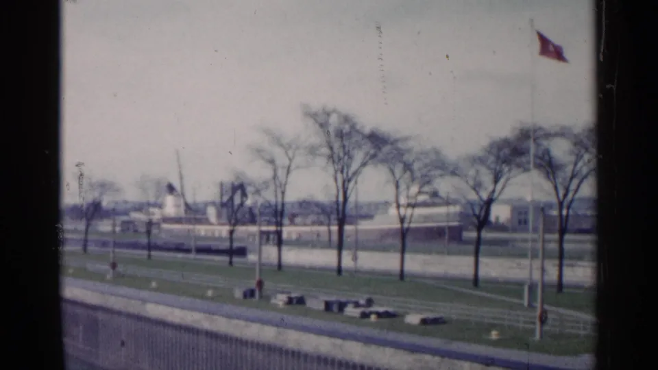 1960 Michigan home-movie frame of bare trees on a grassy waterfront embankment with paved paths and distant industrial buildings.