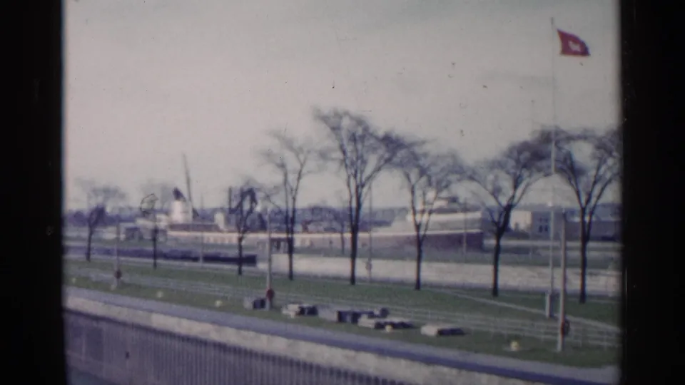Faded 8mm frame from 1960 Michigan showing leafless trees, a red flag, walkways, and distant waterfront industrial structures.