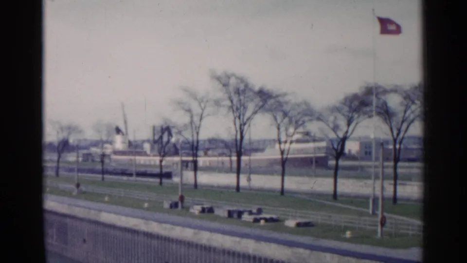 1960 Michigan waterfront view with bare trees, flagpoles, a paved edge, and distant low buildings across the shoreline.