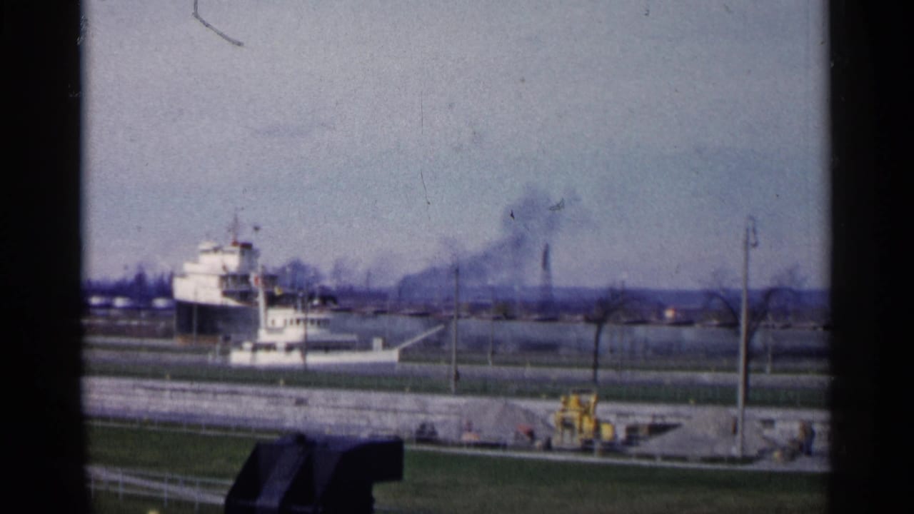 White vessel docked near a grassy waterfront with distant smokestacks and smoke plumes.