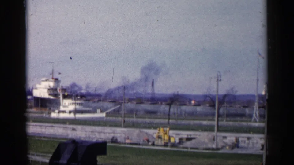 1960 Michigan shoreline view with a docked white vessel, bare trees, distant smoke, and yellow equipment near a paved waterfront area.