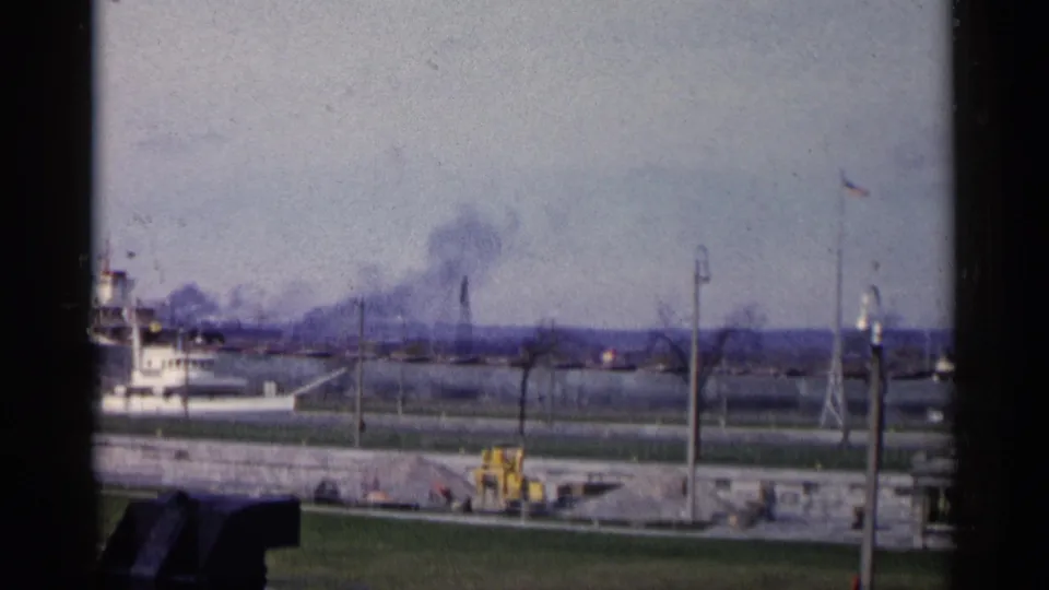 Michigan waterfront frame showing distant smokestacks and smoke, a flagpole, yellow equipment, and the edge of a docked vessel.
