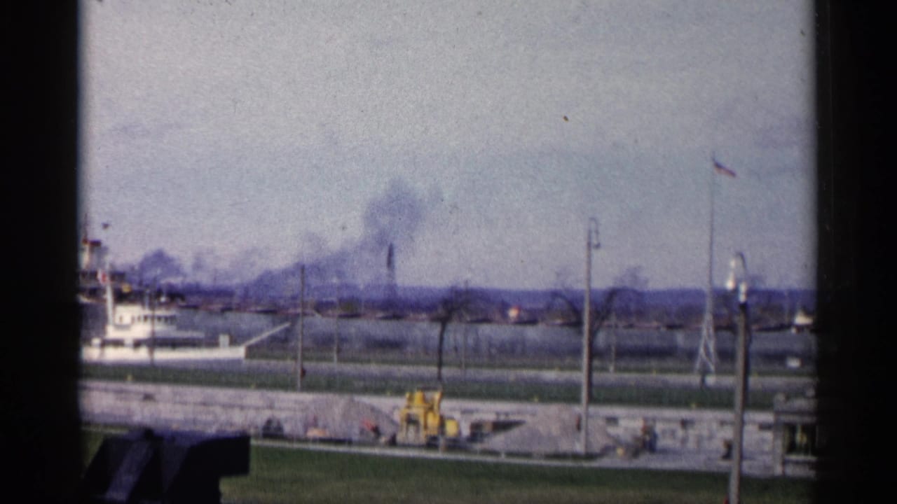 1960 Michigan 8mm frame showing black smoke beyond low industrial buildings, with a yellow tractor, stone wall, grass, and utility poles in the foreground.
